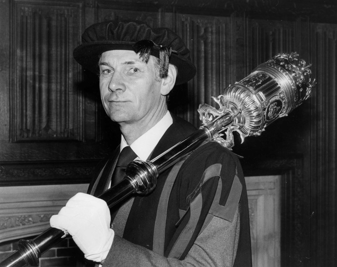 Black and white photo of Dick Smith, he is dressed in an academic gown and a black velvet academic cap. He is carrying a large ceremonial mace that rests on his shoulder.